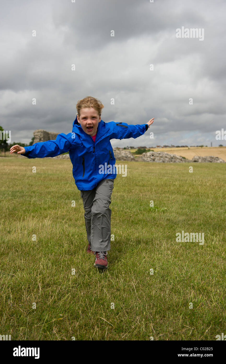 Young girl in outdoor clothing running across a field into a strong wind with her arms outstretched. Stock Photo