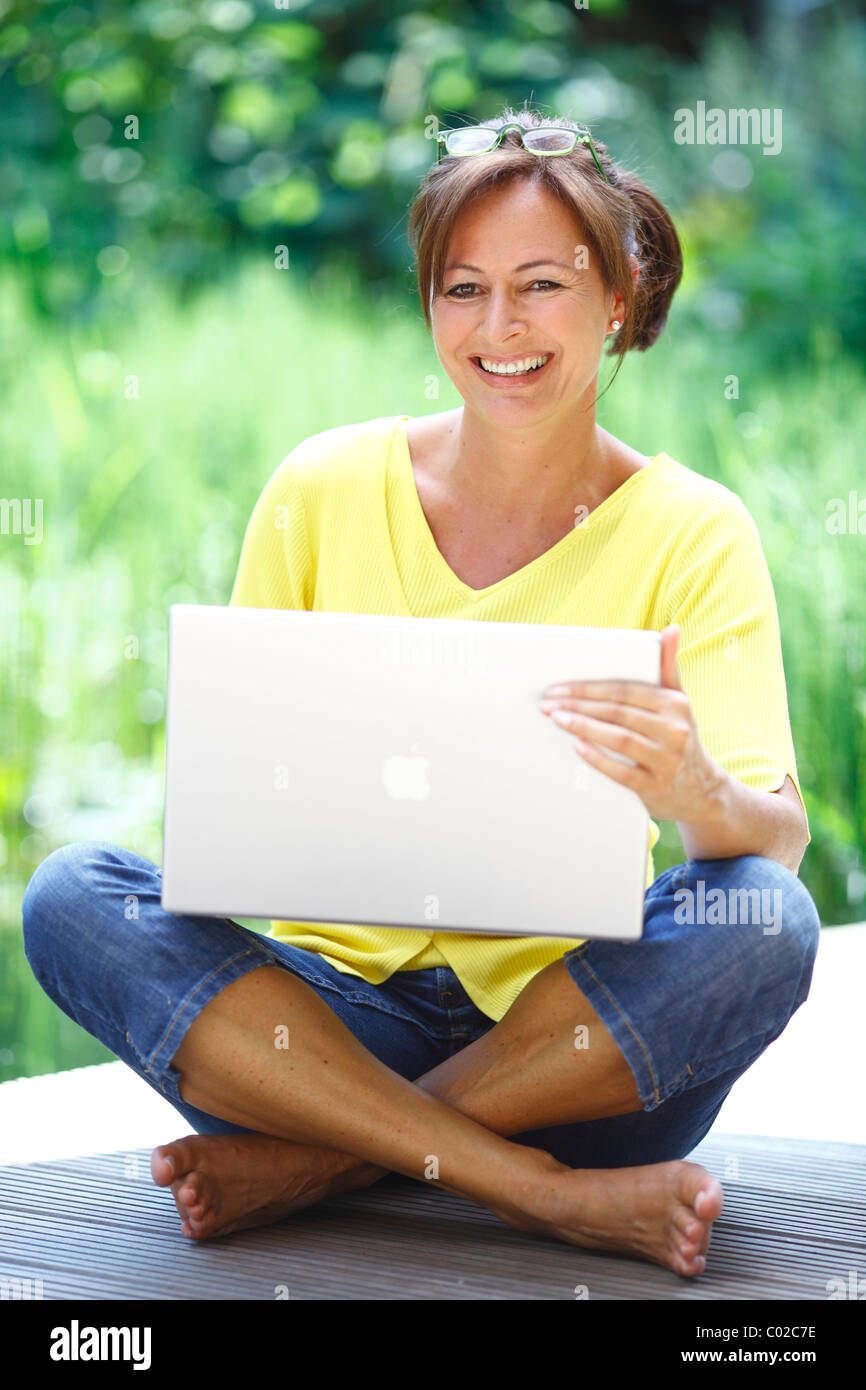 Woman, 45 years, with a Mac Book Pro wearing casual clothes, sitting on a wooden footbridge in natural surroundings Stock Photo