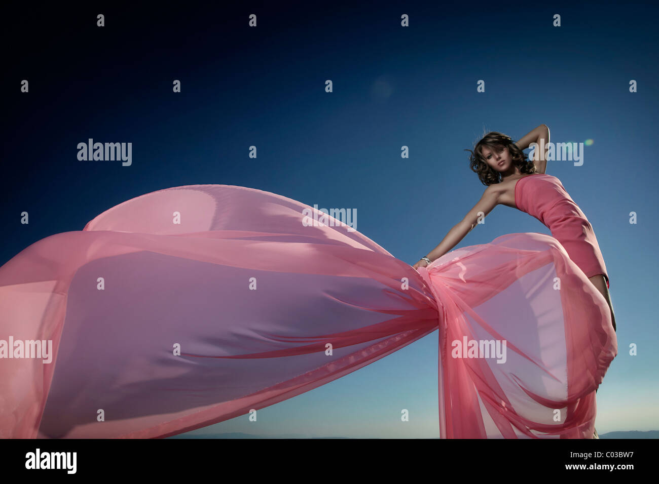 Young woman in a pink dress enveloped in billowing fabric, the setting sun behind her Stock Photo