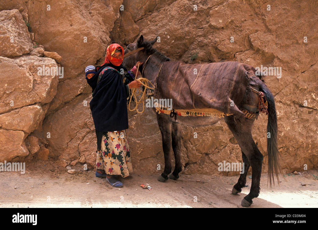 Berber woman in a headscarf and a toddler in a sling holding mule in