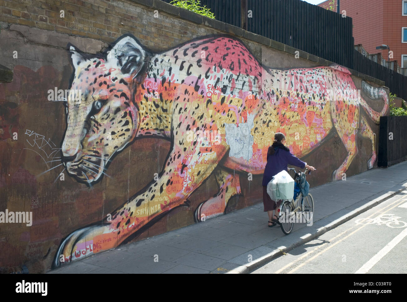 A woman pushing her bicycle up a road, along a wall painted with a pink and yellow leopard, in London Stock Photo