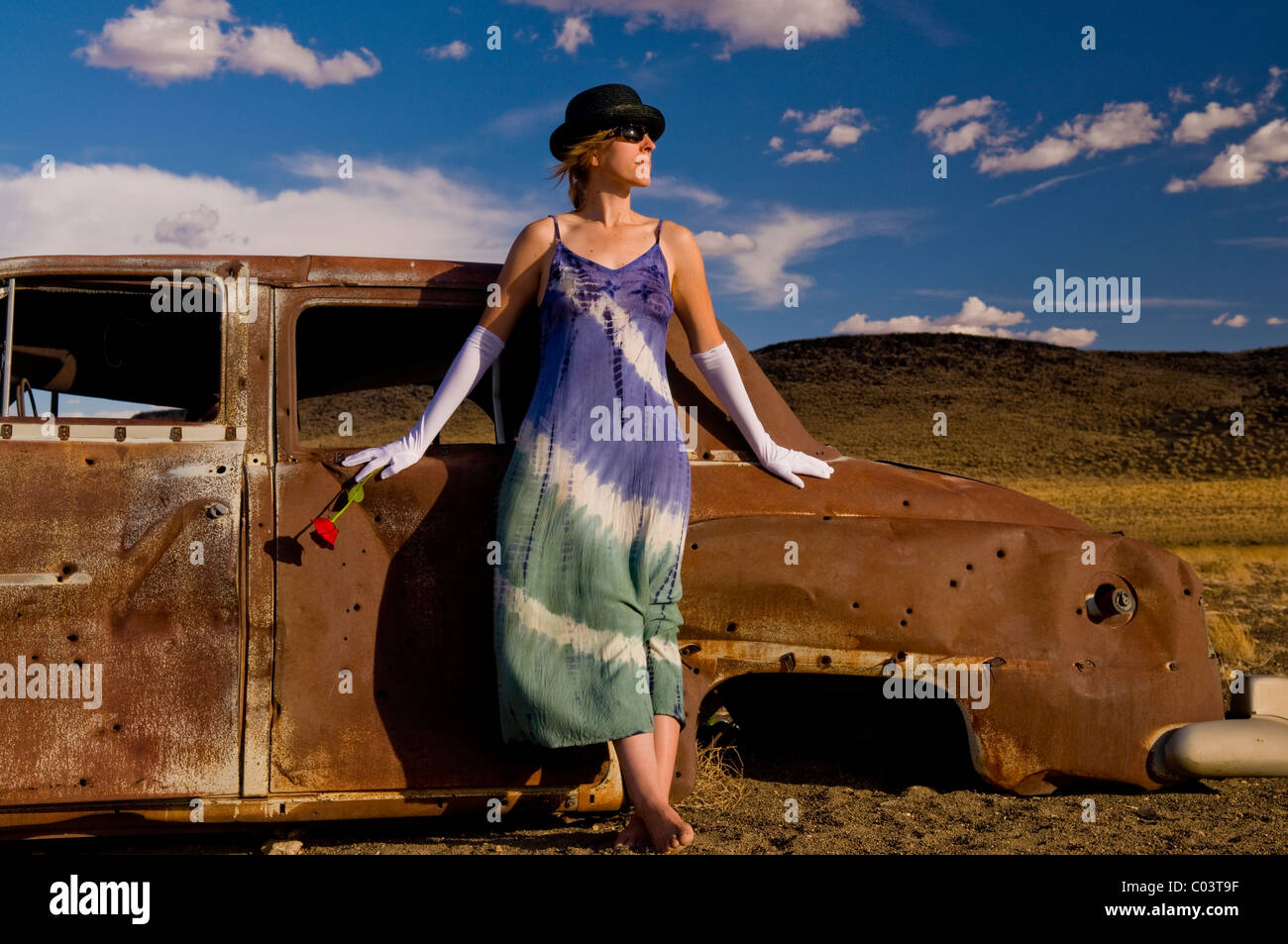 Woman wearing white ballroom gloves, tye died dress, and holding red rose, leans against bullet riddled rusted oid car. Stock Photo
