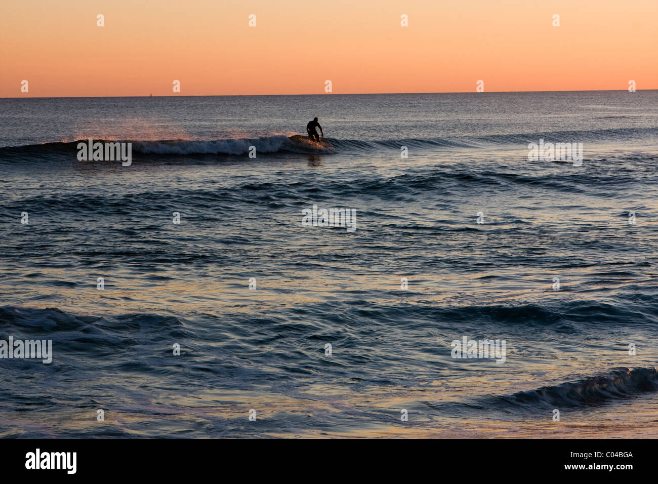 A lone surfer catches the last wave of the evening before sunset. Stock Photo