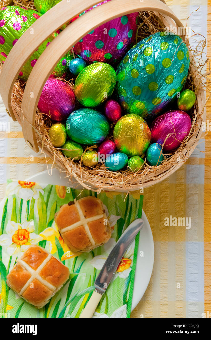 wooden basket filled with colorful foil wrapped chocolate easter eggs on yellow and white tablecloth Hot cross buns on plate Stock Photo