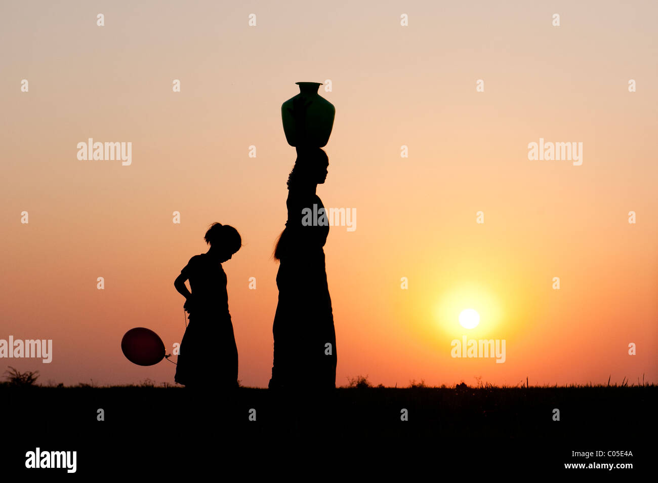 Indian mother carrying water pot with child and balloons in the indian countryside. Silhouette Stock Photo