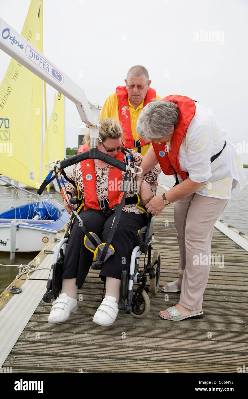 Sailing instructor and helper prepare to lift disabled woman into sailing dinghy using an Oxford Dipper poolside hoist Stock Photo