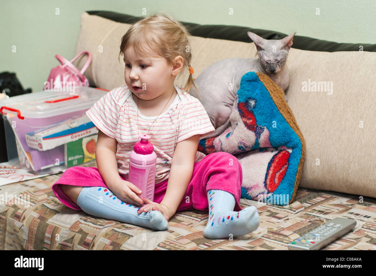Little Russian girl sitting on sofa and naked cat sphinx laying near. Stock Photo