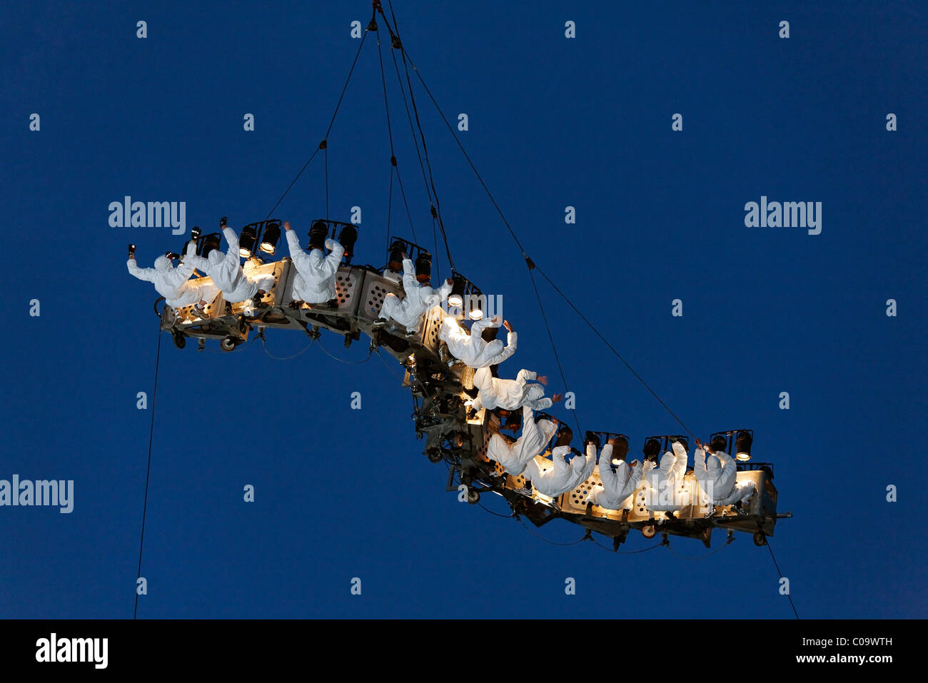White-clad trapeze artists hanging upside down from a waved platform in the air, Global Rheingold, open-air theater by La Fura Stock Photo