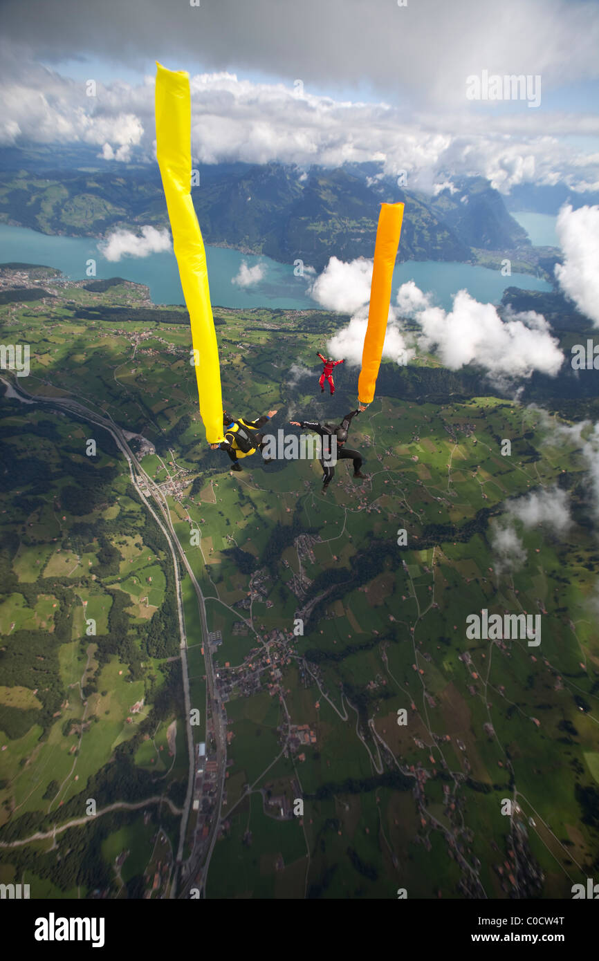 Skydivers with orange and yellow air tubes are falling through the sky together over lakes and green fields. Stock Photo