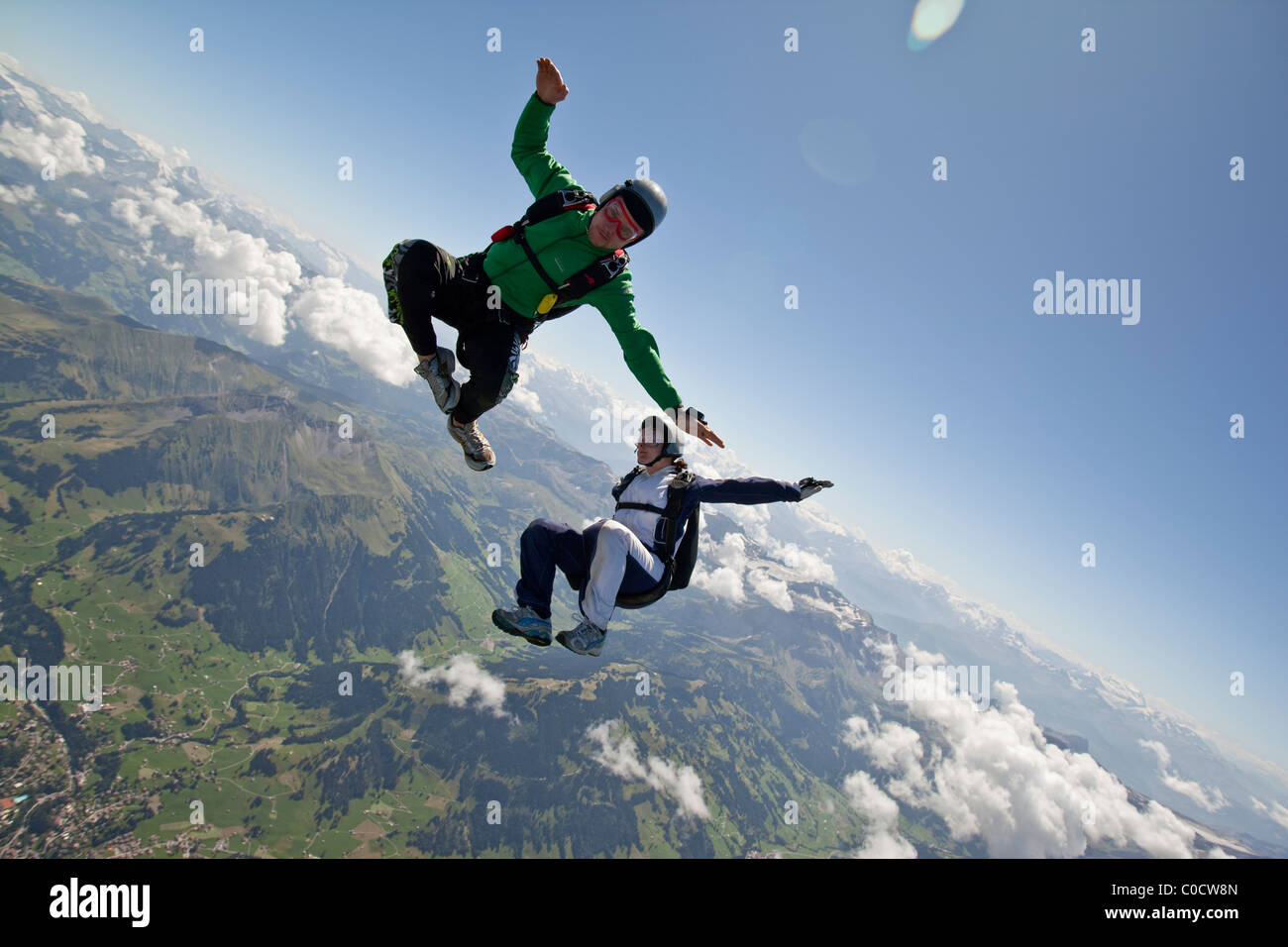 Man in green jumpsuit and girl in white dress are skydiving together over cloudy mountains area. Stock Photo