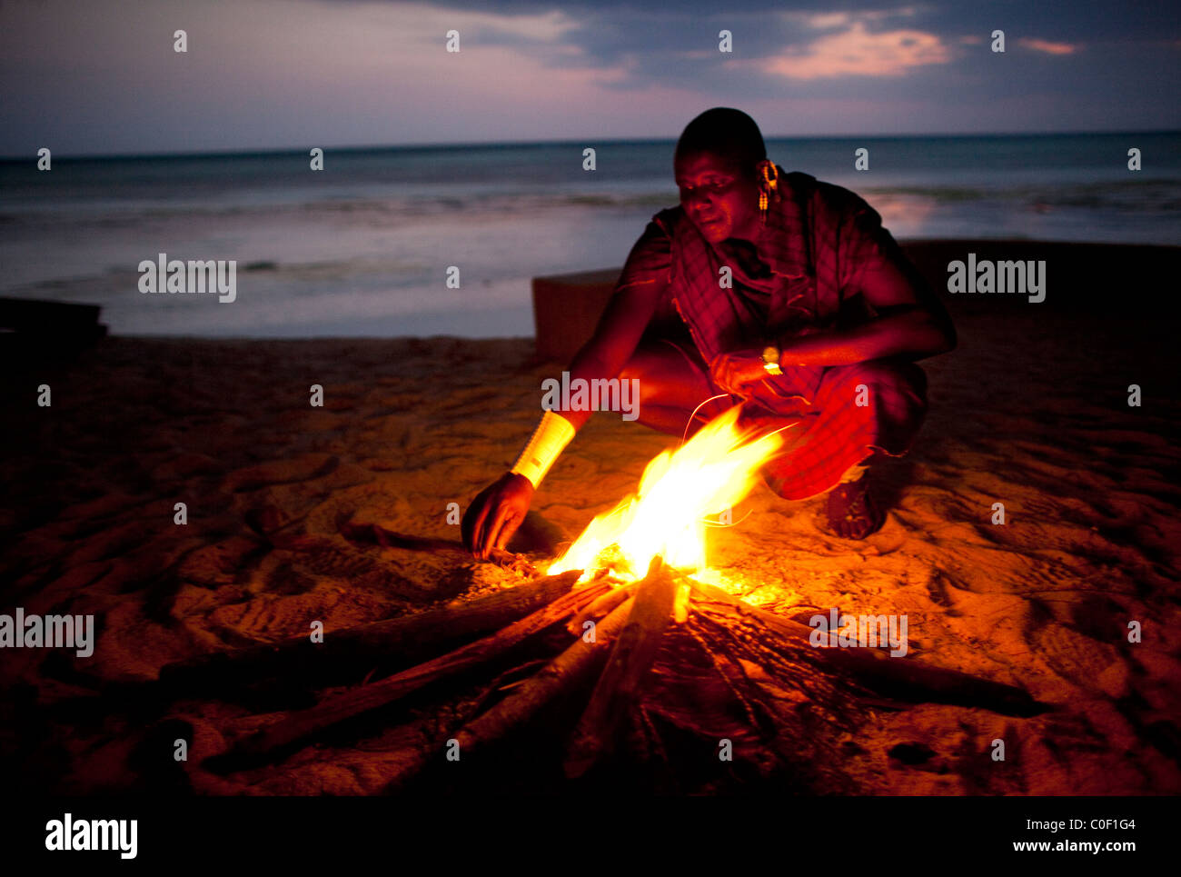 A member of  the Maasai tribe in northern Tanzania, working as a watchmen for local hotels, tends to a bon fire on the beach. Stock Photo