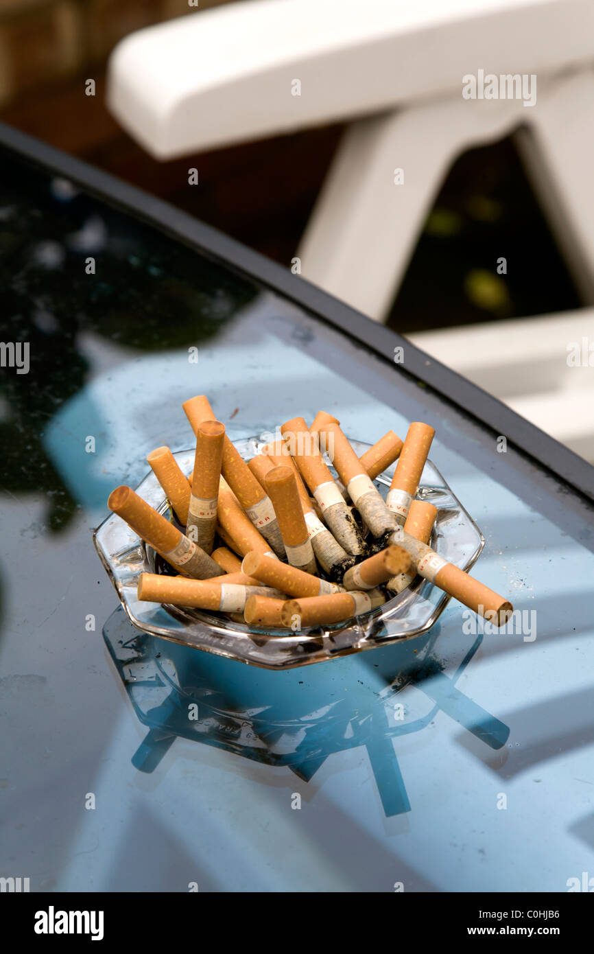 cigarette garden A FULL ASHTRAY OF CIGARETTE BUTTS ON A GLASS TABLE IN THE GARDEN Stock  Photo