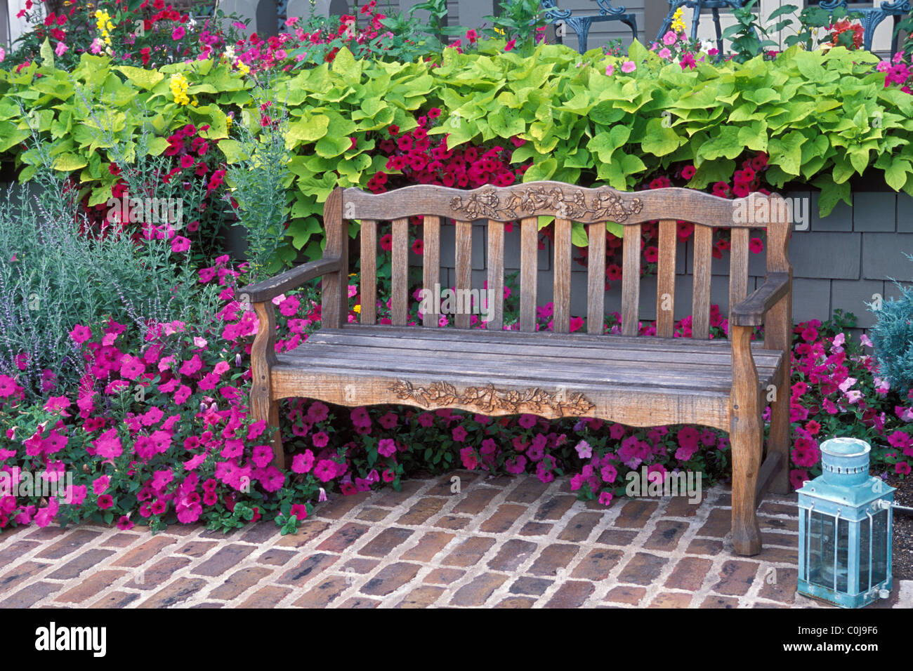 GARDEN BENCH ON BRICK PATIO BORDERED BY ORNAMENTAL SWEET POTATO VINE, RUSSIAN SAGE AND PETUNIAS.  CANDLE LANTERN IN CORNER. Stock Photo
