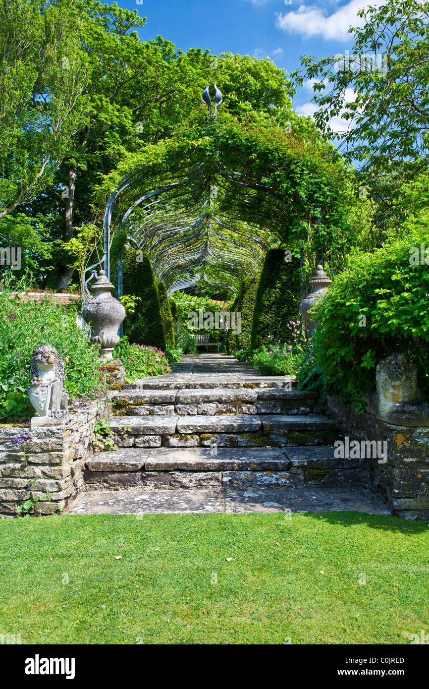 Stone steps leading to a pergola over a paved path in an English country garden with a wooden seat at the end. Stock Photo