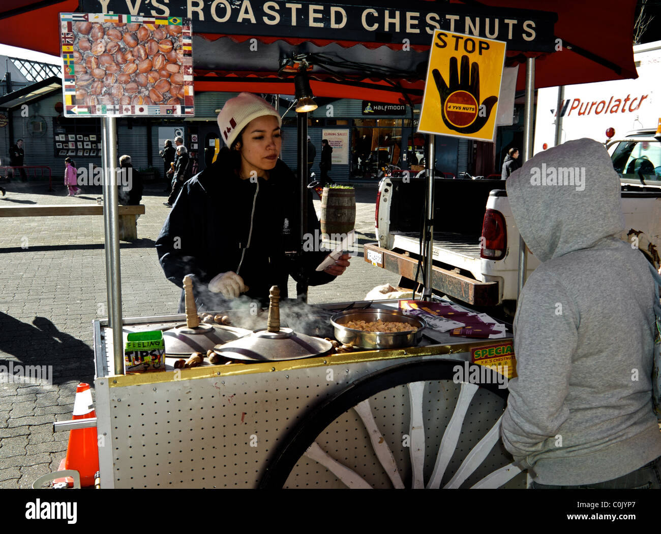 Young woman selling hot roasted chestnuts pistachios from cart at entrance to Granville Island Public Market Vancouver Stock Photo