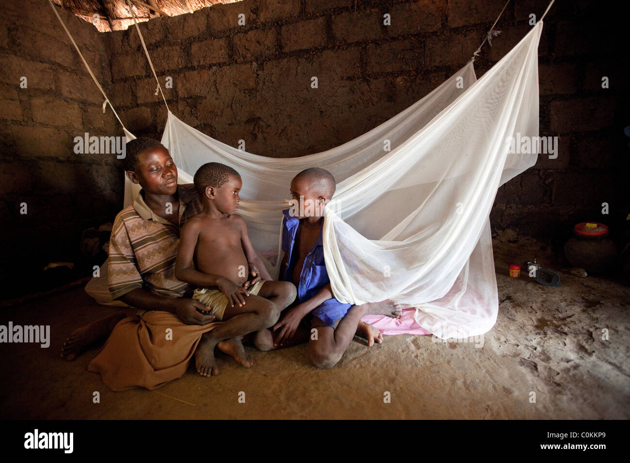 A single mother sits with her children outside the family's bed, draped with a mosquito net. Amuria, Uganda, East Africa. Stock Photo