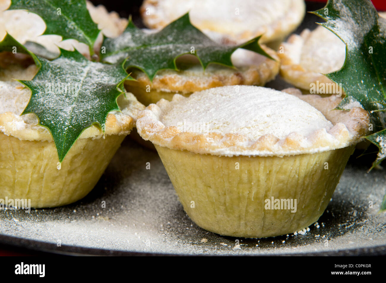 Close up of mince pies dusted with icing sugar on black plate with holly sprigs Stock Photo