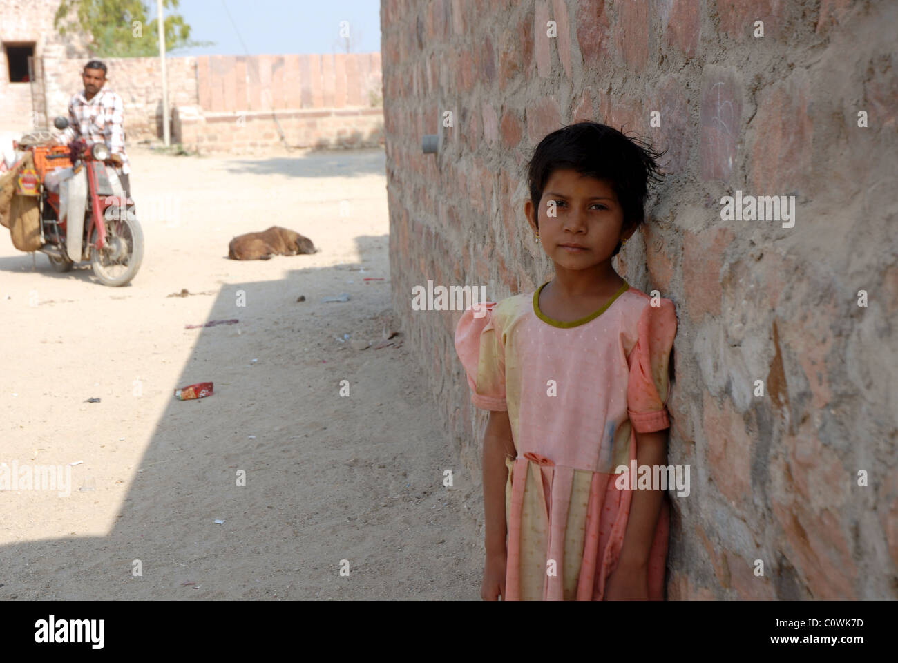 portrait of girl leaning of wall in Rajasthan with father and sleeping dog in background Stock Photo
