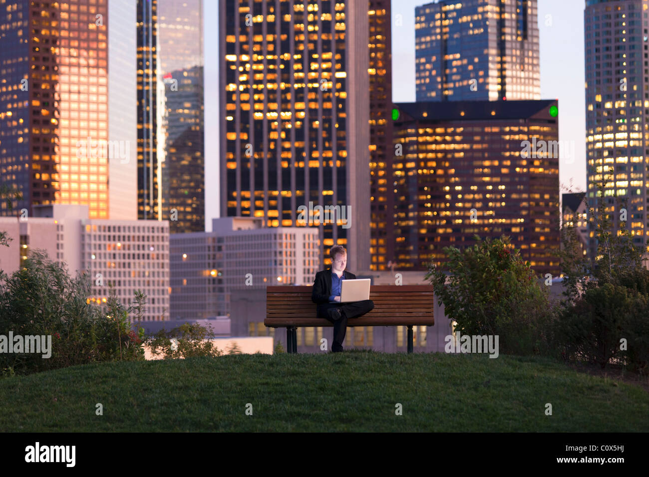 Male sitting on park bench working on laptop computer with Los Angeles city skyline in background at dusk, sunset Stock Photo