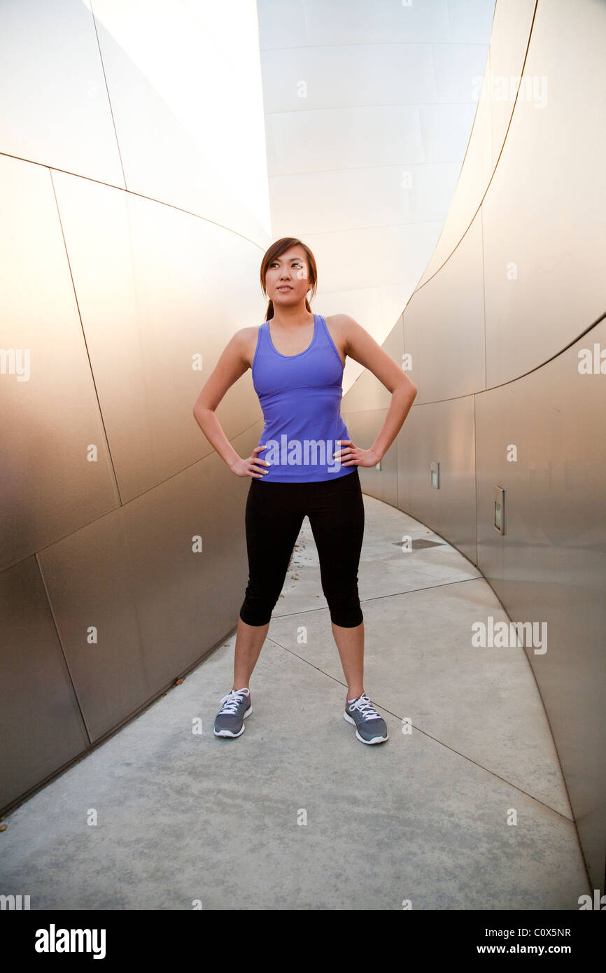 Asian American female runner resting with hands on hips standing in front of metal clad wall.  Purple tank top and black pants. Stock Photo