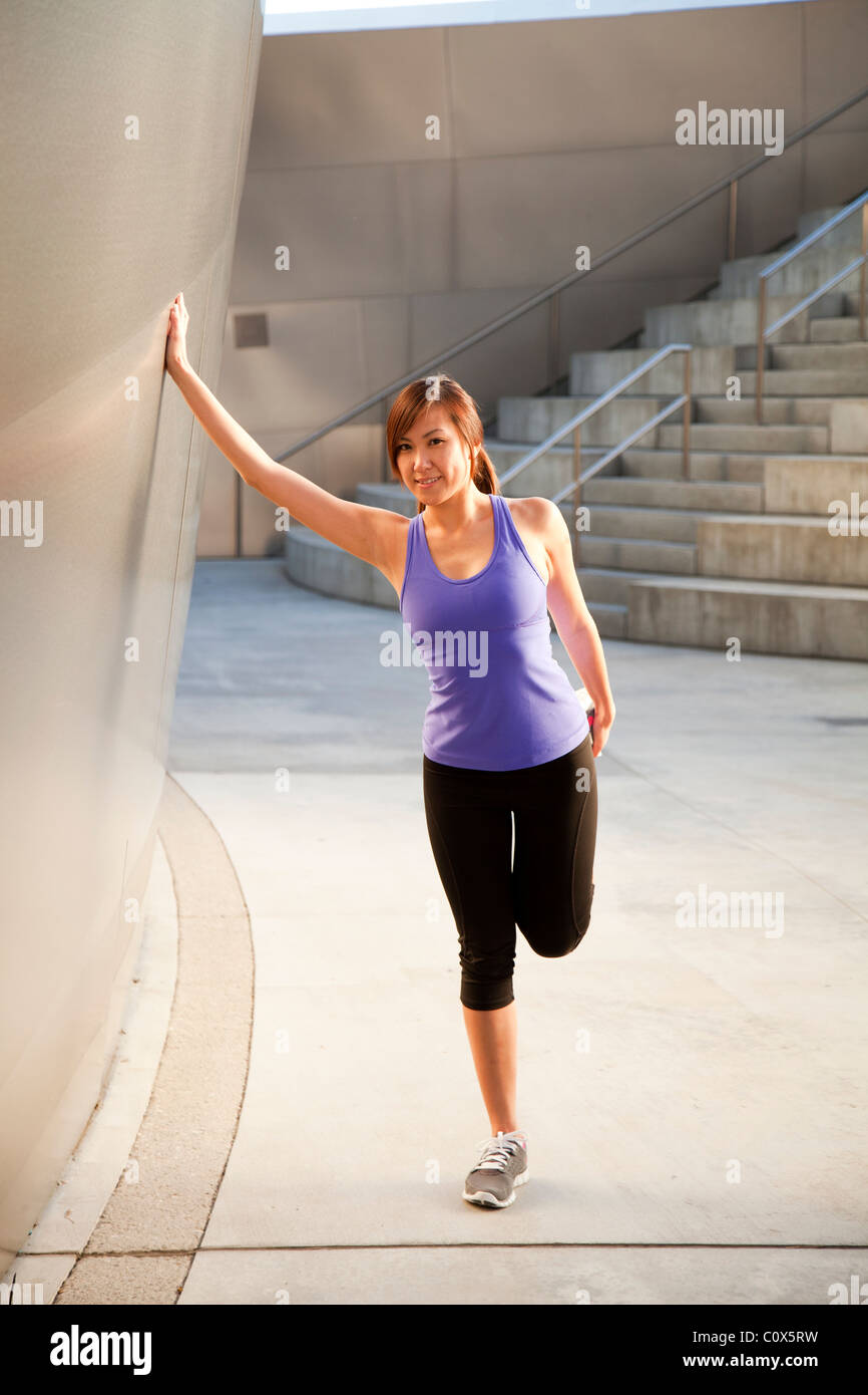 Asian American female runner stretching while leaning against metal clad building.  She is wearing purple tank top. Stock Photo