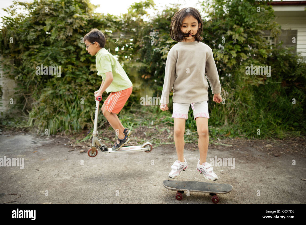 Girl jumps on skateboard as boy bunny-hops on scooter. Stock Photo