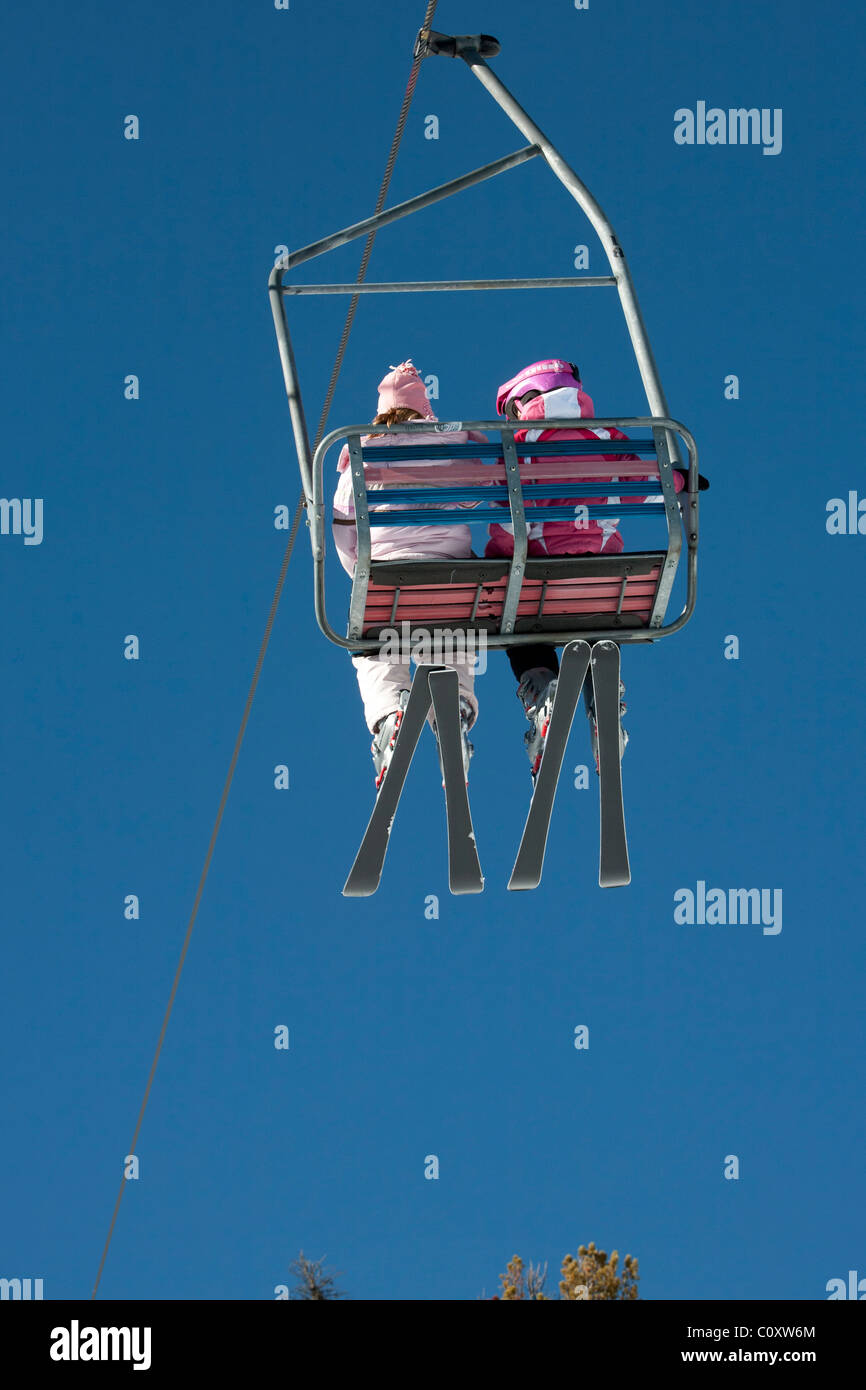 Brightly clothed young skiers ride a chair lift on a sunny day. Skiers are framed by blue sky. Stock Photo
