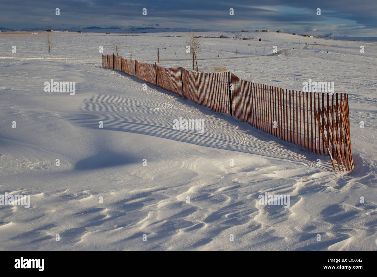 Rough scallops in drifted snow at the end of a fence and smooth drifts behind the snow fence show its impact on wind patterns. Stock Photo