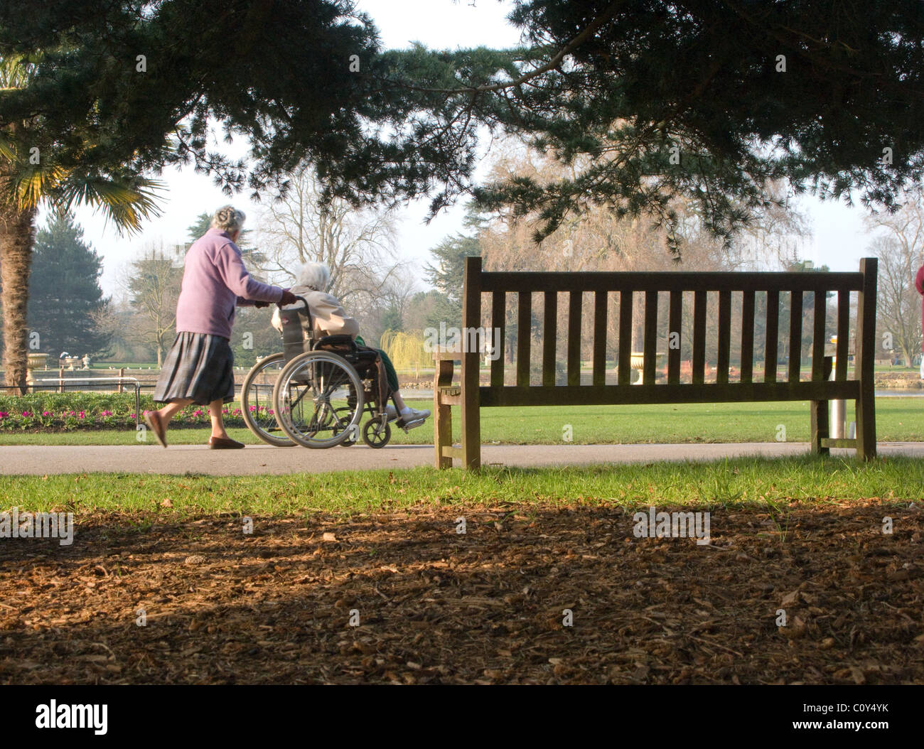 A woman pushes a wheelchair past a park bench in Kew Gardens, London. The gardens are particularly well suited for wheelchairs Stock Photo
