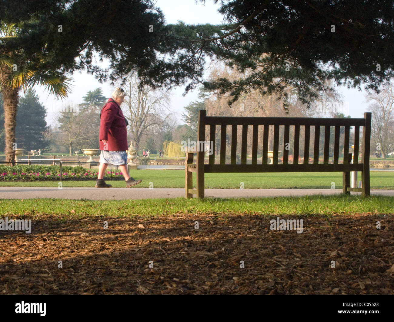 A slightly eccentric looking woman walking past a park bench in Kew Gardens, London Stock Photo