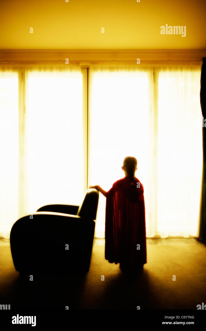 Girl in red cape stands at window beside armchair Stock Photo