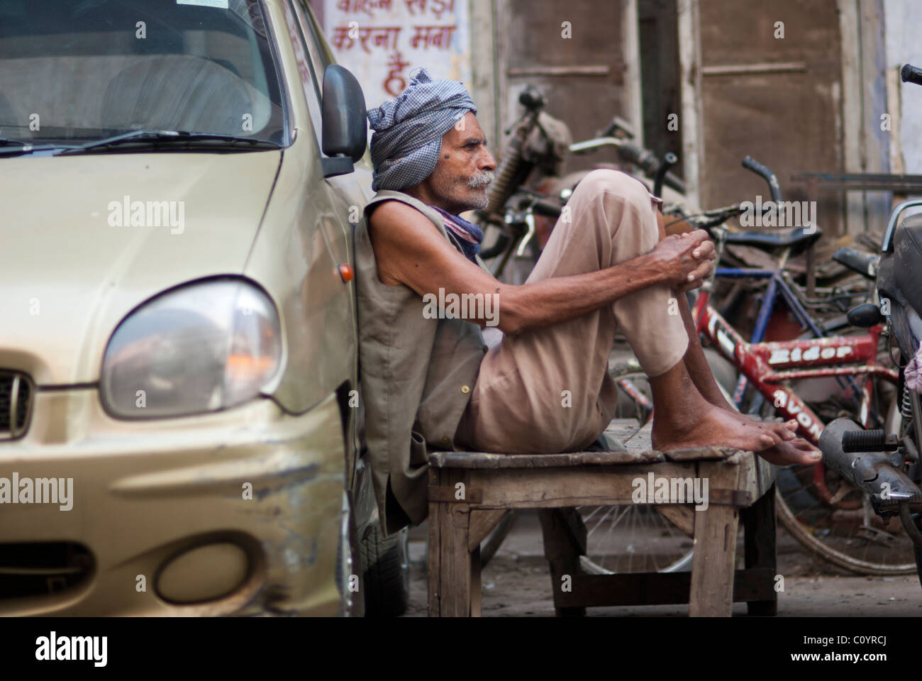 Indian man sitting on a stool on a street leaning against a car Stock Photo