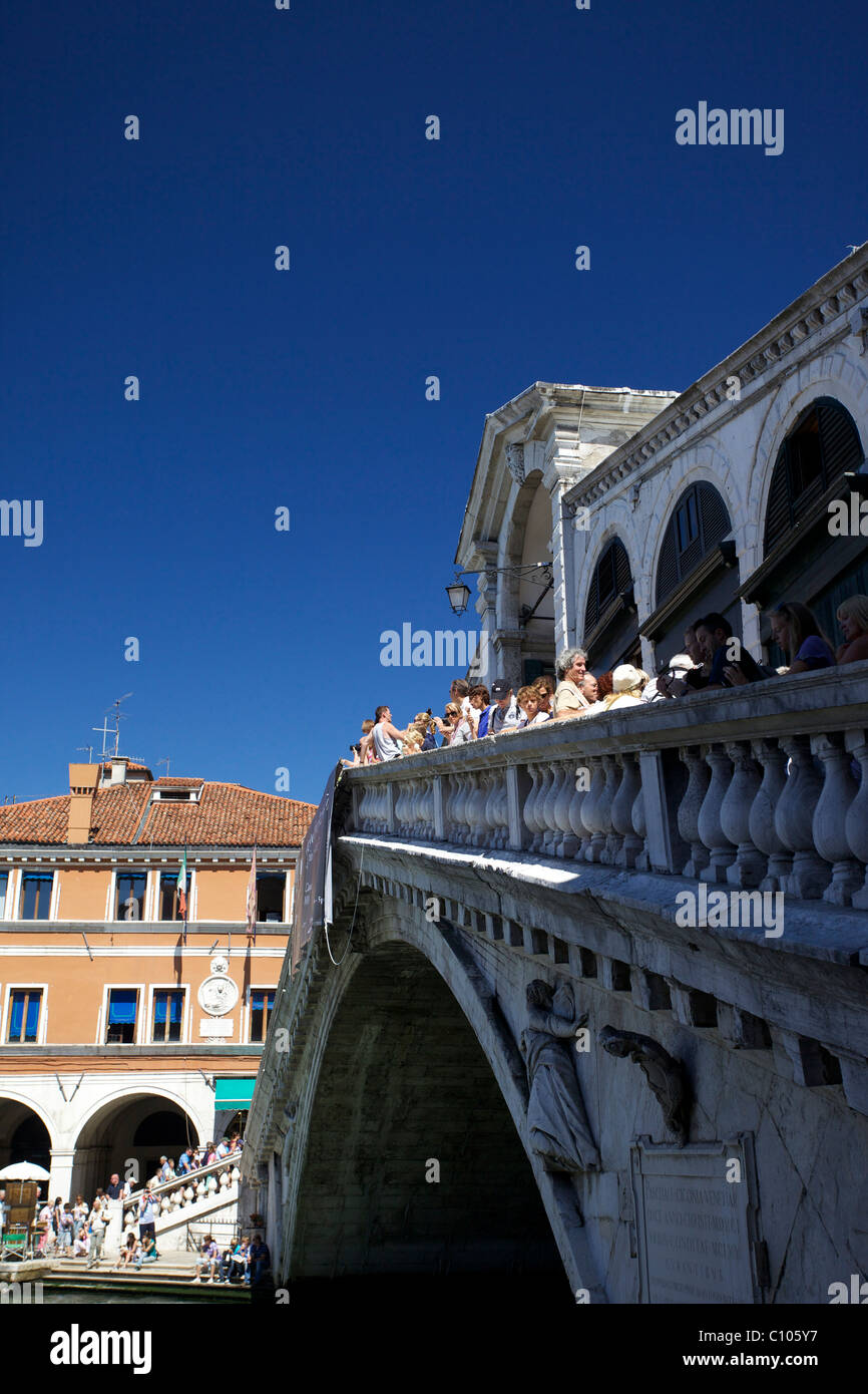 The Ponte Rialto bridge in Venice Italy as seen from the side with tourists lookng up river in bright sunshine and blue skies. Stock Photo