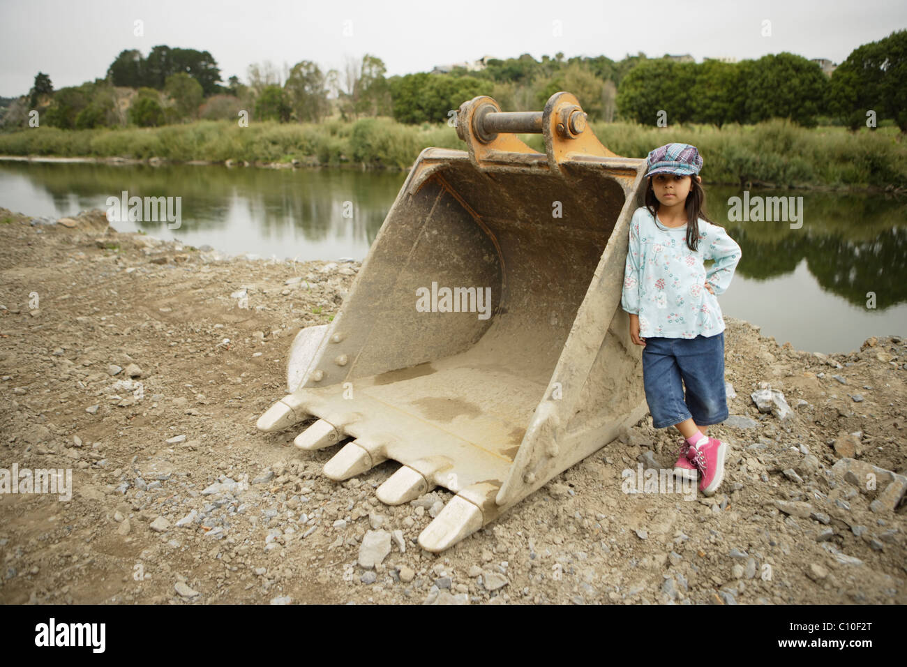 Five year old girl leans against digger shovel at the construction of flood defences, Manawatu river, Palmerston North, NZ Stock Photo