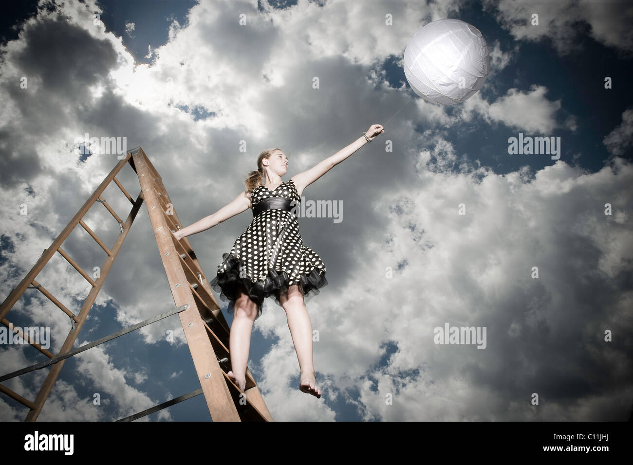 A teenage girl wearing a black and white polka dotted dress stands on a ladder while holding a white ball. Stock Photo
