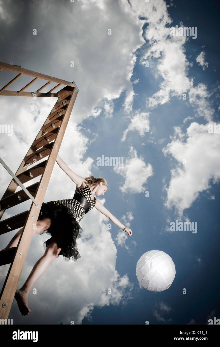 A teenage girl wearing a black and white polka dotted dress stands on a ladder while holding a white ball. Stock Photo
