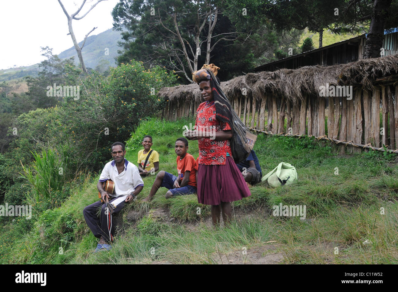 Dani woman coming from the field while young Dani people sing Christian songs with guitar accompaniment, Kurima, Baliem Valley Stock Photo