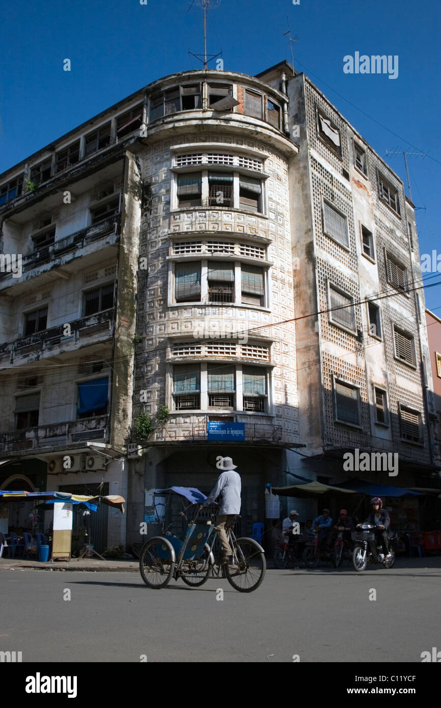 A male cyclo driver is peddling a vehicle on a city street past a bright but rundown apartment building in Phnom Penh, Cambodia. Stock Photo