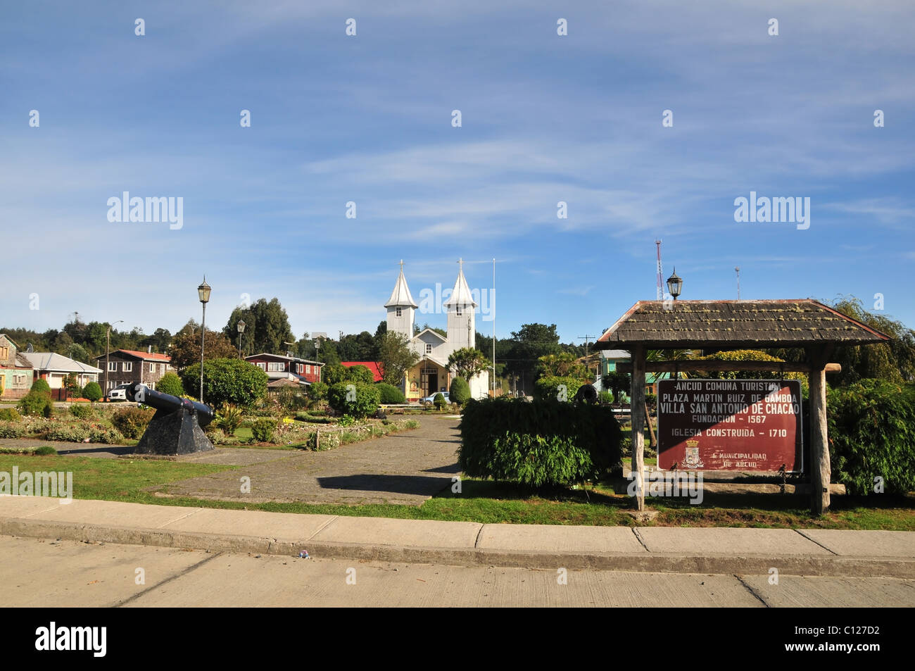 Blue sky view of Plaza Martin Ruiz, with information sign, black cannon and silver church of San Antonio, Chacao, Chiloe, Chile Stock Photo