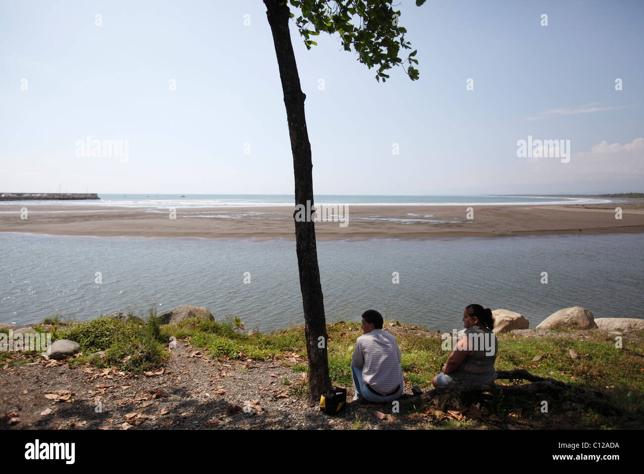 A man and a woman sit in the shade of a tree overlooking the beach and the entrance to the harbor in Quepos, Costa Rica Stock Photo