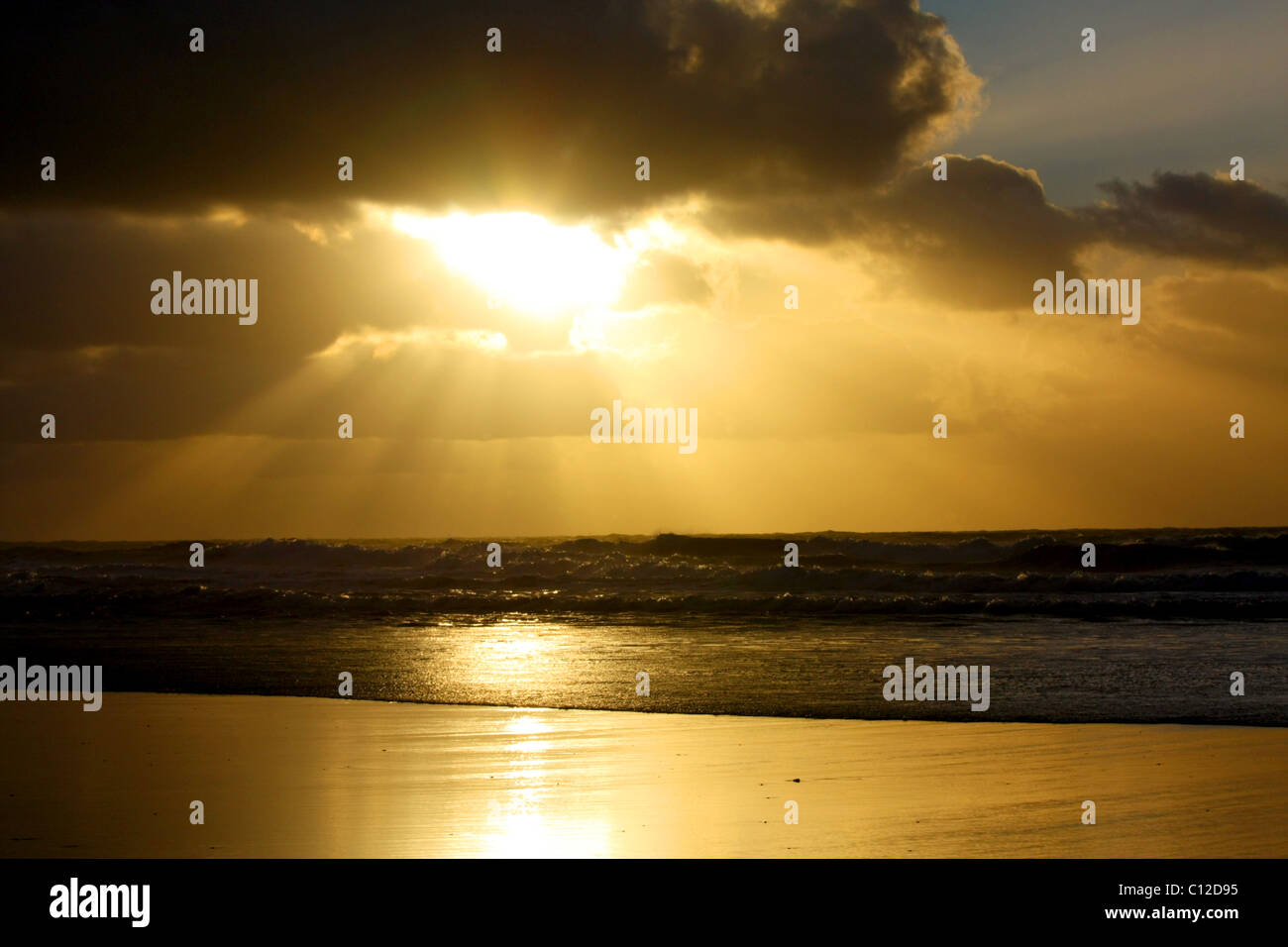 40,151.06066a Sun rays sunbeams dark clouds cloud layers golden glow shining sunlight onto ocean waves beach. Stock Photo