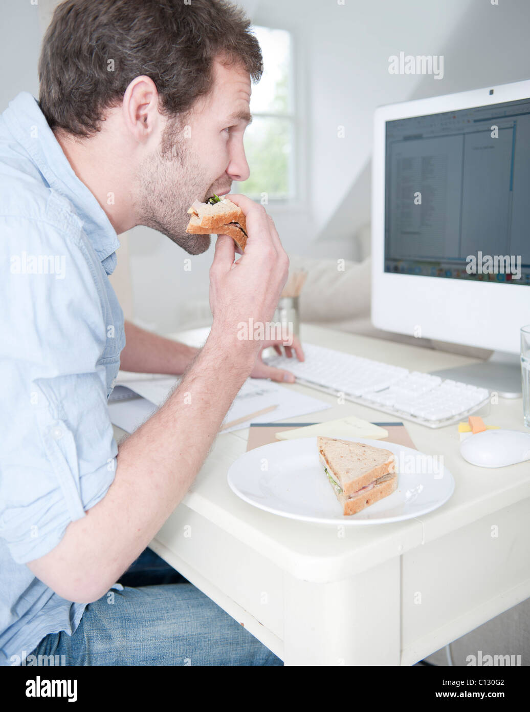 young man at desk eating sandwich and working with computer Stock Photo