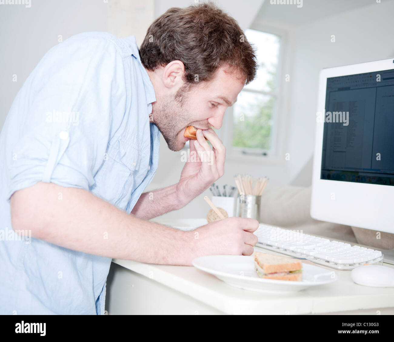 young man at desk eating sandwich and working with computer Stock Photo