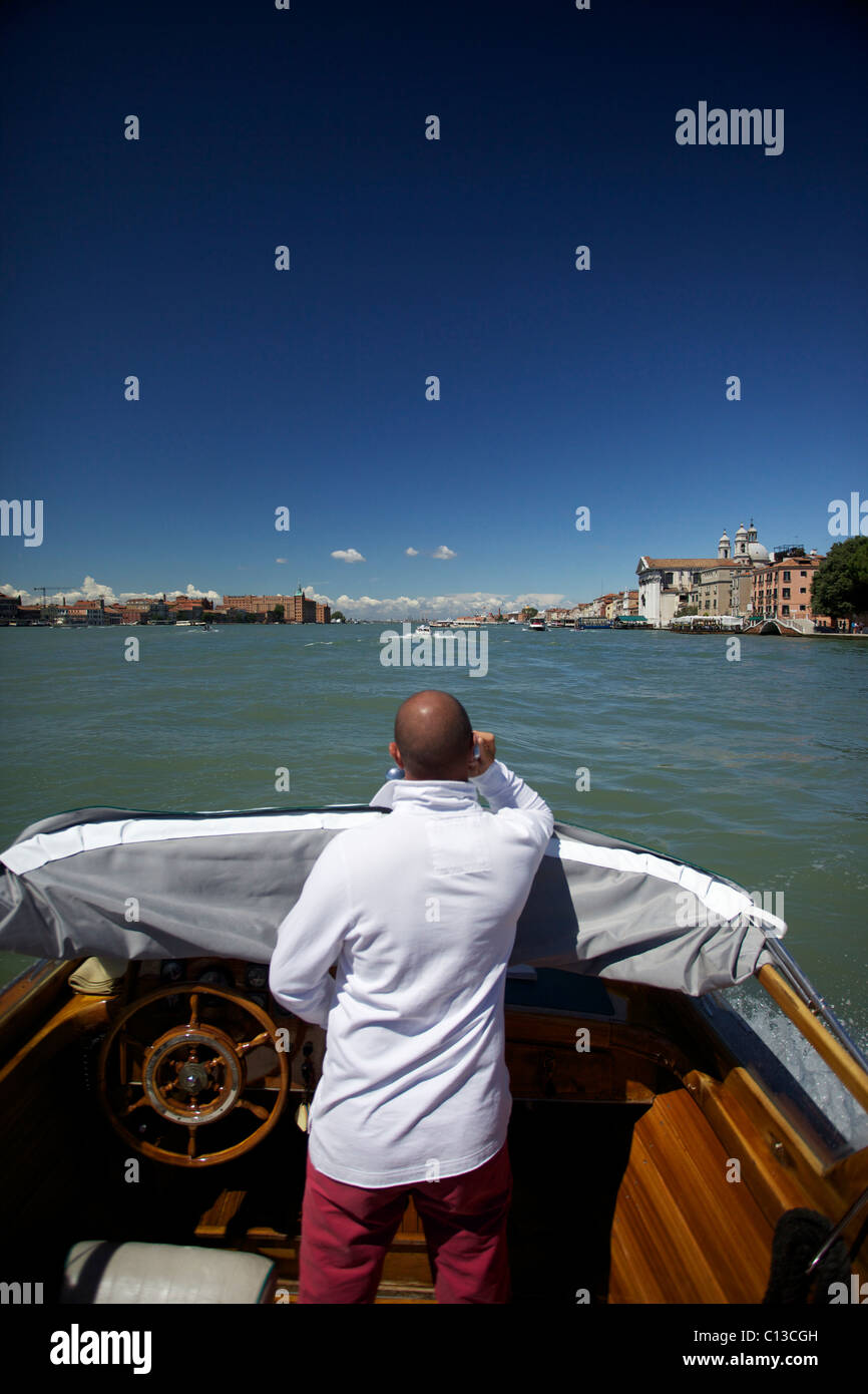 A driver/sailor/pilot of a water taxi speaks on mobile phone whilst steering a private water taxi in Venice Italy Stock Photo
