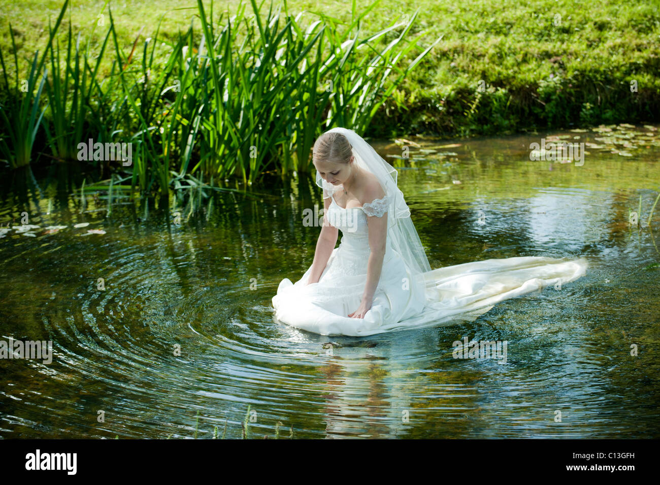 woman lady walking into lake water up to her waist in long wedding dress with lily pads and reeds Stock Photo