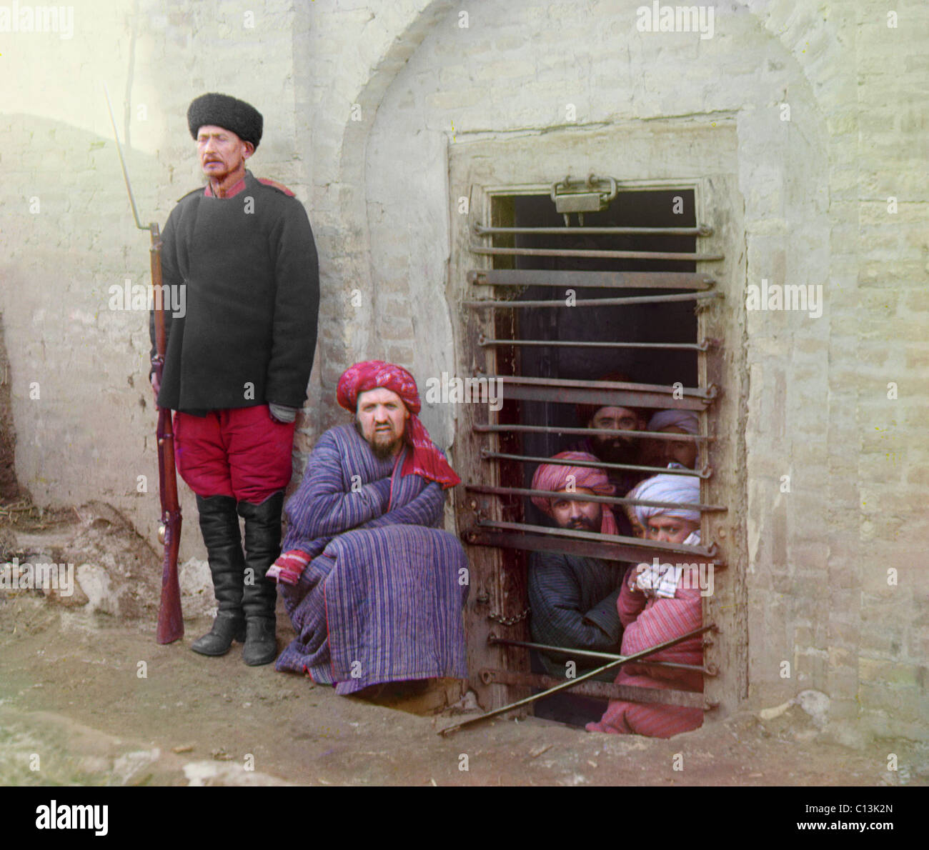 Traditional Central Asian prison, with cells cut into the ground. Turbaned Inmates looking out through the bars while guard stands by with a Russian rifle, uniform, and boots. Russian Empire, ca. 1910. Color photo by the Czar's photographer, Sergei Prokudin-Gorskii, Stock Photo