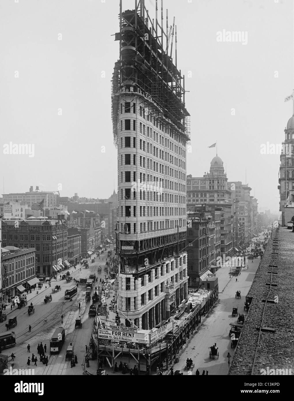 Flatiron Building during construction in 1902. The steel frame construction, a rectangular grid of I-beams, ascends above the stone facing. Signs advertise space for rent in what was then called the 'Fuller Building.' Stock Photo