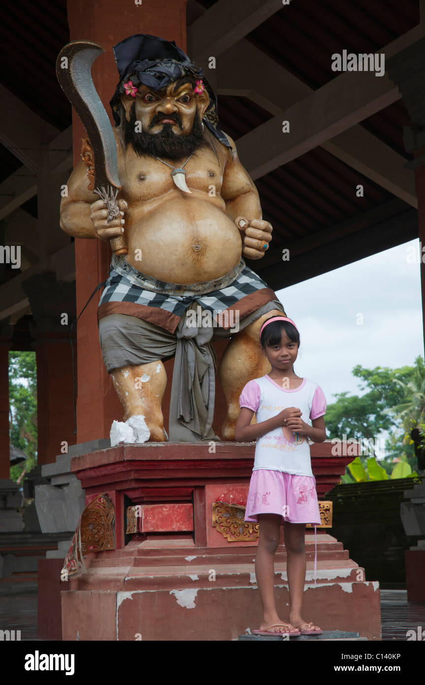 A young girl standing in front of a fiercely grotesque temple sculpture in Ubud, Bali, Indonesia Stock Photo