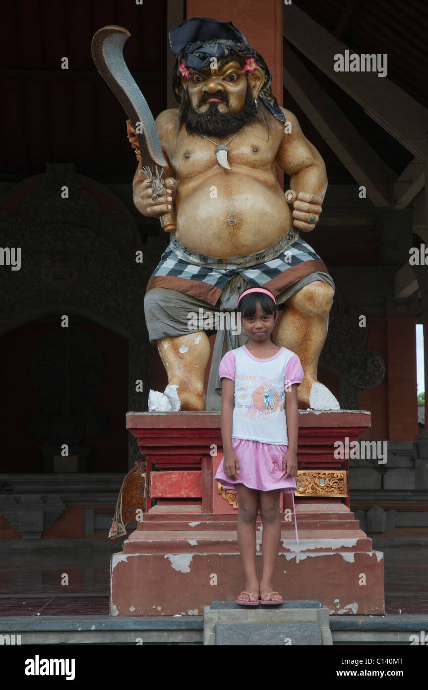 A young girl standing in front of a fiercely grotesque temple sculpture in Ubud, Bali, Indonesia Stock Photo