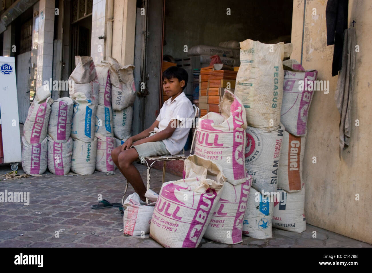 A young child laborer boy is sitting among several cement sacks on a city sidewalk in Phnom Penh, Cambodia. Stock Photo